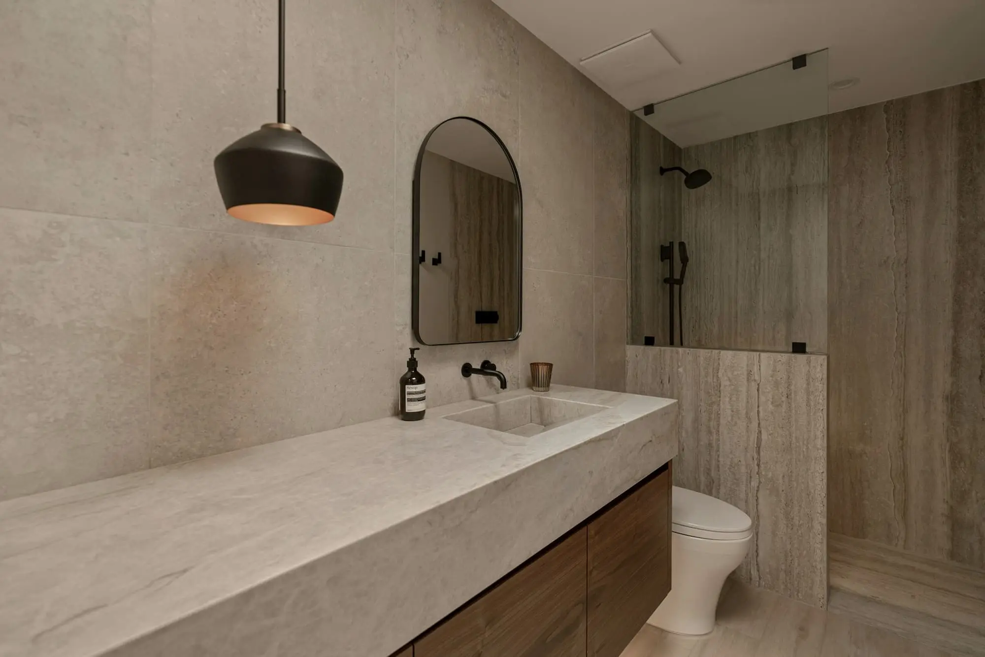 Long stone vanity, arched mirror and a walk-in shower with travertine walls
