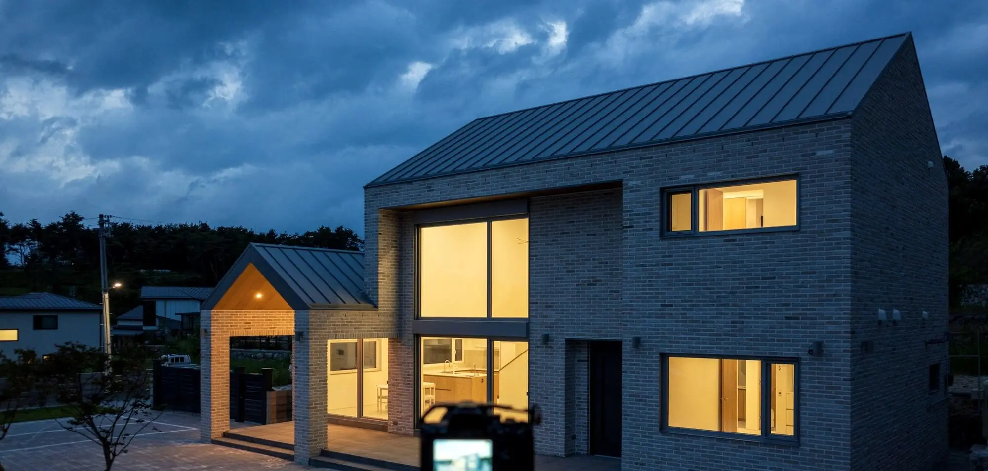 Grey brick house with a zinc roof and a double-height window, glowing at dusk