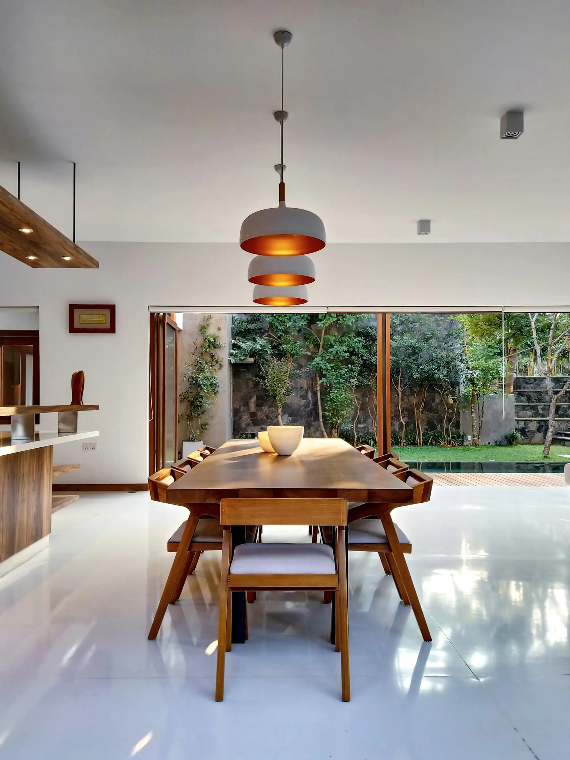 Dining room with a long walnut table beneath copper pendants, opening onto a garden